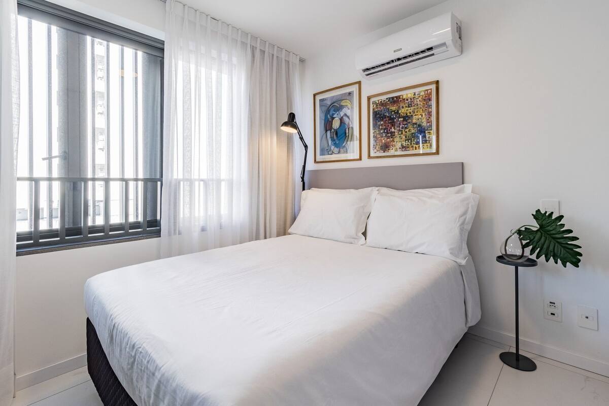 Habitación de hotel con cama, ventana con persianas, aire acondicionado, dos cuadros enmarcados, lámpara de pie y planta en maceta.