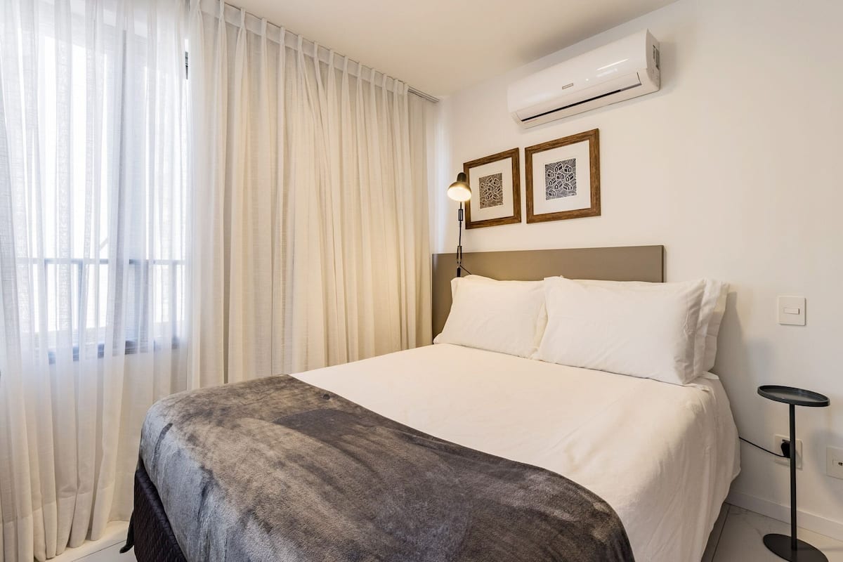 Una habitación de hotel con una cama, aire acondicionado, una lámpara y dos cuadros en la pared.