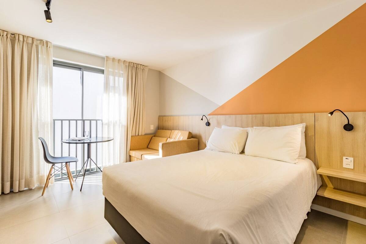 Una habitación de hotel con cama, una mesita con silla, un sofá y un balcón con cortinas.
