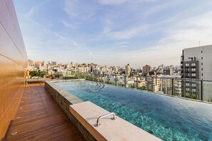 Piscina infinita en la azotea con vista a la ciudad, terraza de madera y barandillas metálicas.