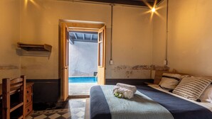 Una habitación con una cama, una silla de madera, piso a cuadros y vista a una piscina por una puerta abierta.