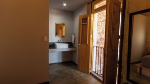 Un baño con lavamanos, espejo y puerta de madera que da a un balcón con pared de ladrillo.