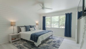 Un dormitorio con cama, mesitas de noche, ventilador de techo, ventana con cortinas y vistas al exterior.