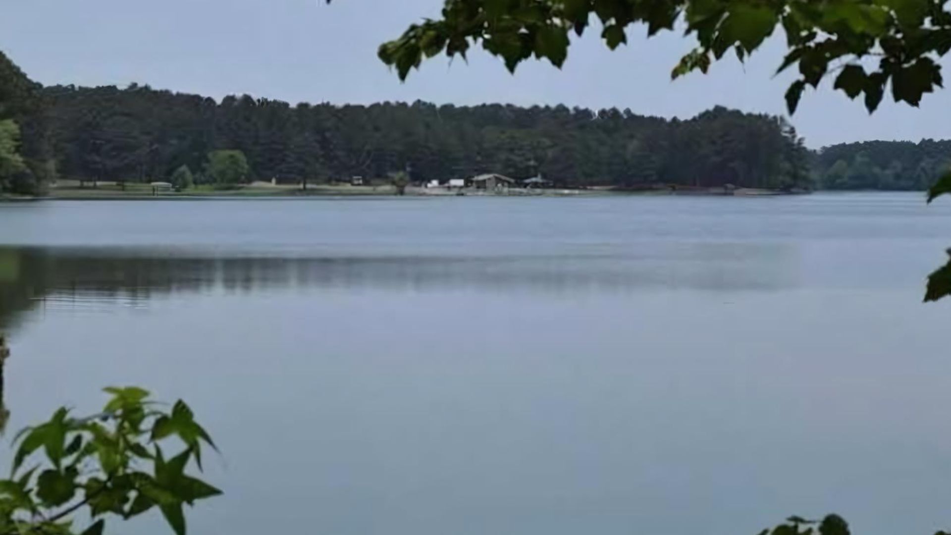 A calm lake with a forested shoreline and a few buildings in the distance.