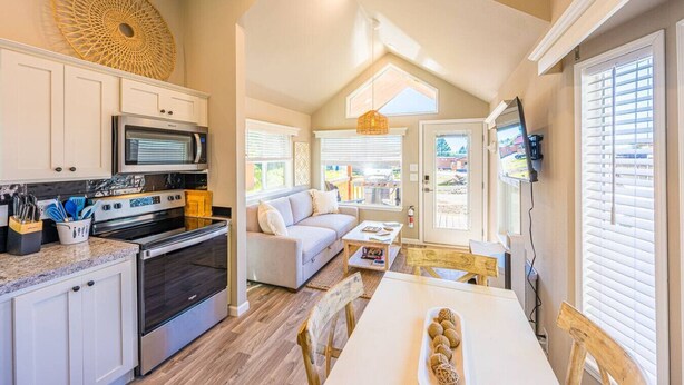 A modern kitchen with white cabinets, stainless steel appliances, and a dining area with a white table and wooden chairs. - Rainforest Tiny Home Retreat: Coastal (Forks)