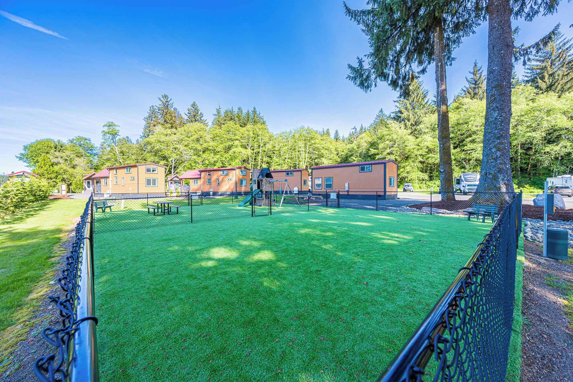 A fenced outdoor area with a green lawn, a playground, and a row of cabins.