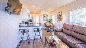 A modern living room with a brown leather sofa, a coffee bar with stools, a dining area with a wooden table, and a kitchen with a flat-screen TV mounted on the wall.