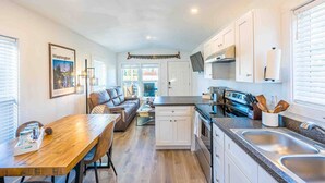 A compact kitchen with white cabinets, a dining area with a wooden table and chairs, and a view of the outdoors through the windows.