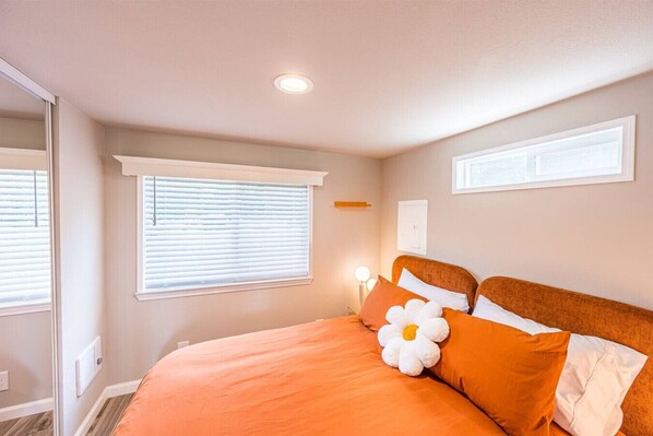 Une chambre avec un grand lit, deux fenêtres équipées de stores, un luminaire au plafond et un thermostat fixé au mur.