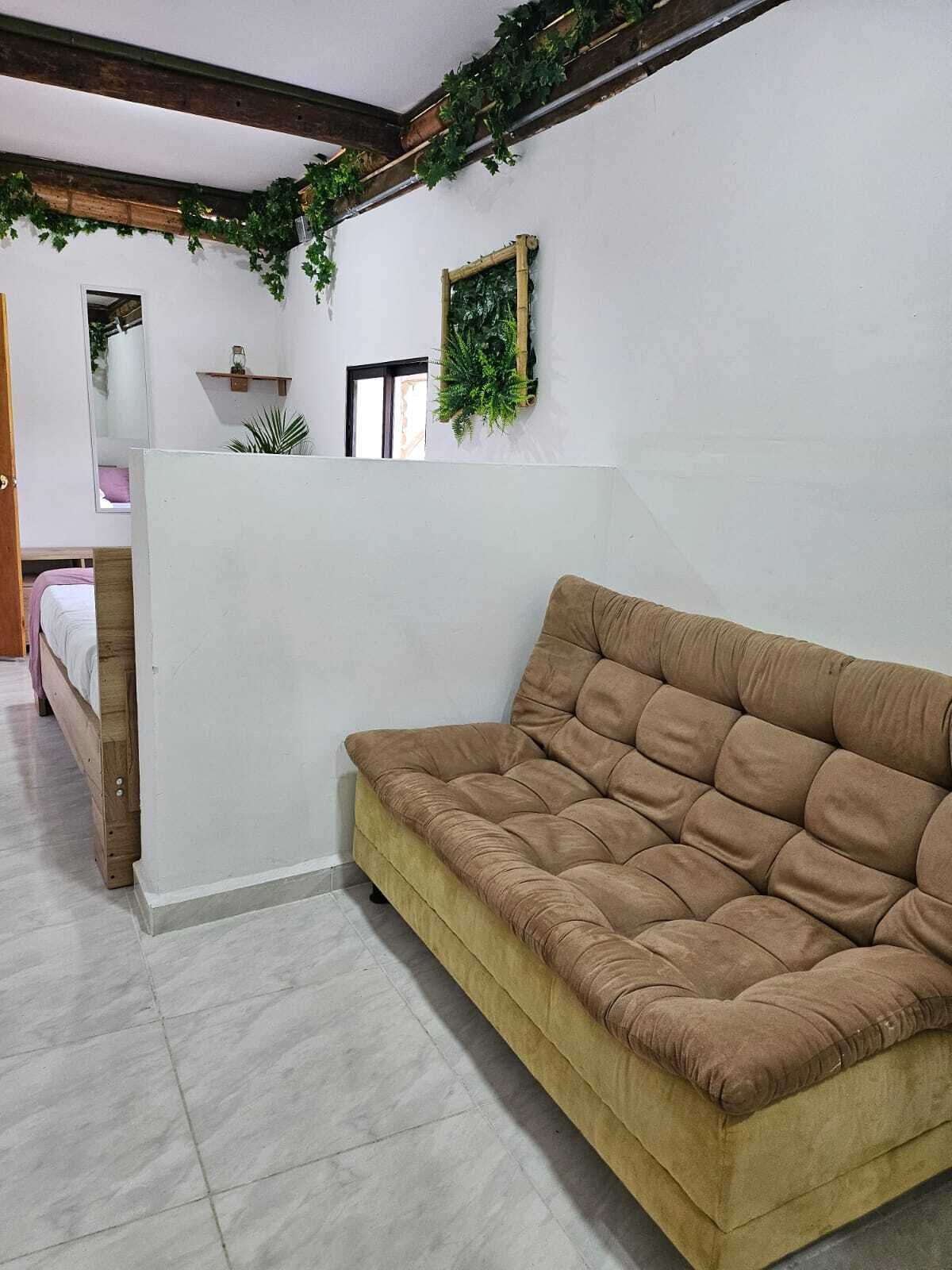 Una habitación con un sofá marrón, una cama con cabecera de madera, un separador blanco y una ventana con vistas al exterior.