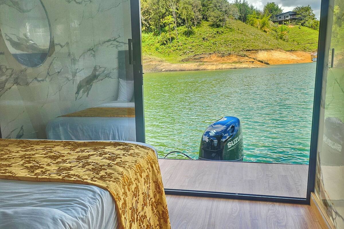 A room with a bed, a view of a body of water, and a boat in the distance.