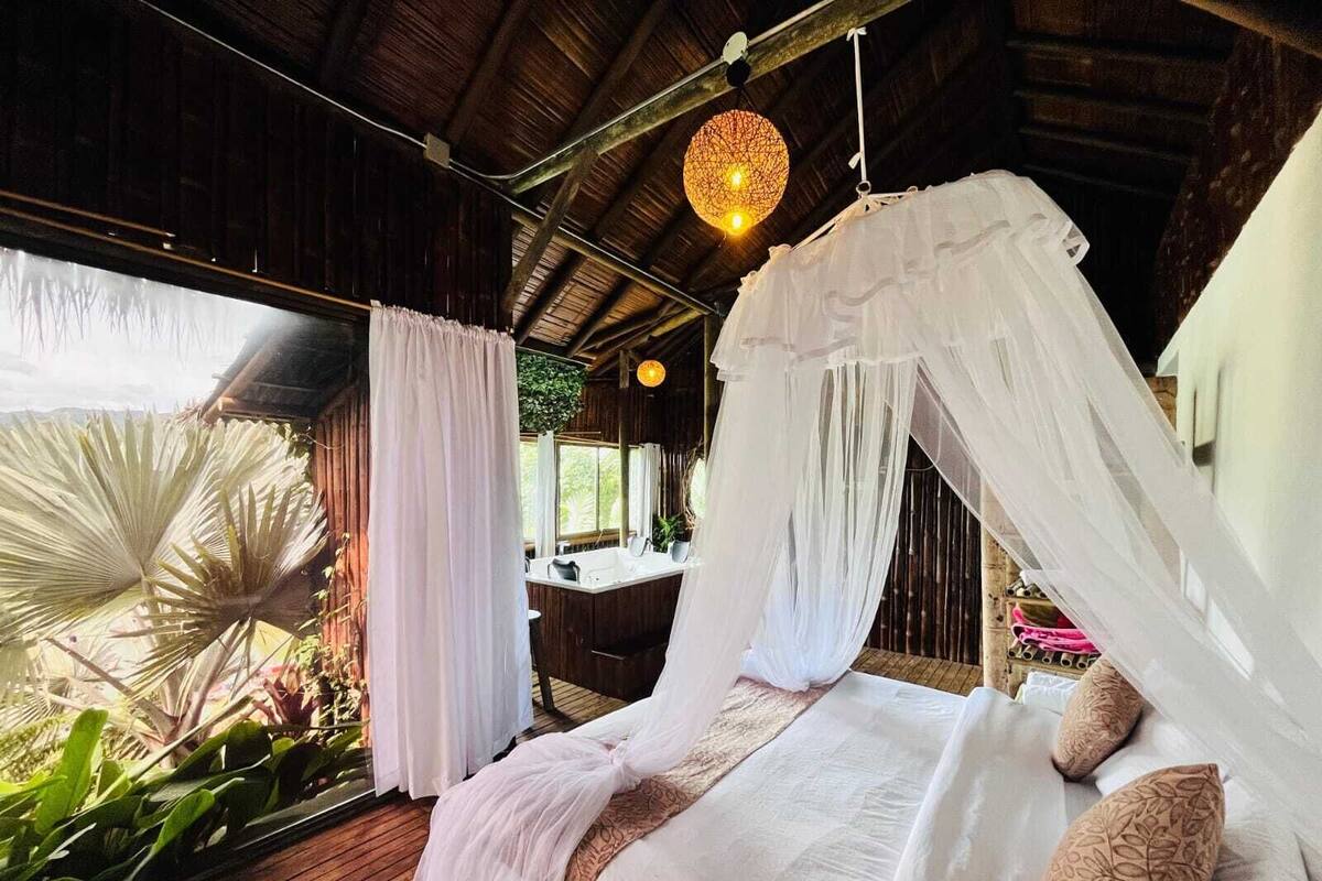 A bedroom with a canopy bed, a wooden floor, and a view of greenery outside.