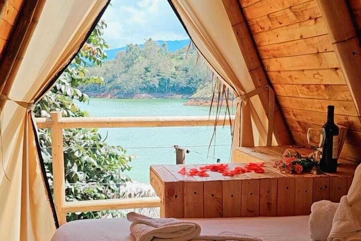 A cozy wooden tent with a bed, a table with a bottle and glass, and a view of a lake and mountains.