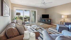A living room with a sofa, coffee table, and TV, a ceiling fan, and a view through a glass door to an outdoor area. - Quaint Oceanfront, Pets ok, Pools, Golf, Amenity card! 1389 Pelican Watch (Seabrook Island)