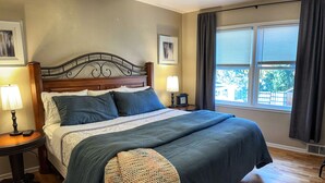 Ein Schlafzimmer mit einem großen Bett, einem hölzernen Kopfteil, zwei Nachttischen mit Lampen, einem Fenster mit Vorhängen und Blick ins Grüne.