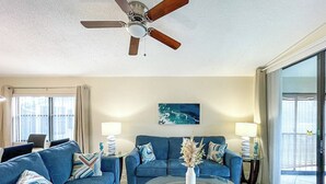Ein Wohnzimmer mit zwei blauen Sofas, einem Glastisch, einem Deckenventilator und einem großen Fenster mit Vorhängen.
