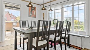 Un espace repas comprenant une grande table, six chaises, des luminaires suspendus et de grandes fenêtres donnant sur l’extérieur.
