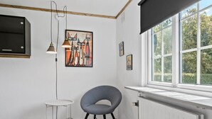 Une pièce avec un radiateur blanc, une chaise grise, une petite table blanche et un meuble noir. Il y a une fenêtre donnant sur des arbres et un tableau représentant des chats accroché au mur.