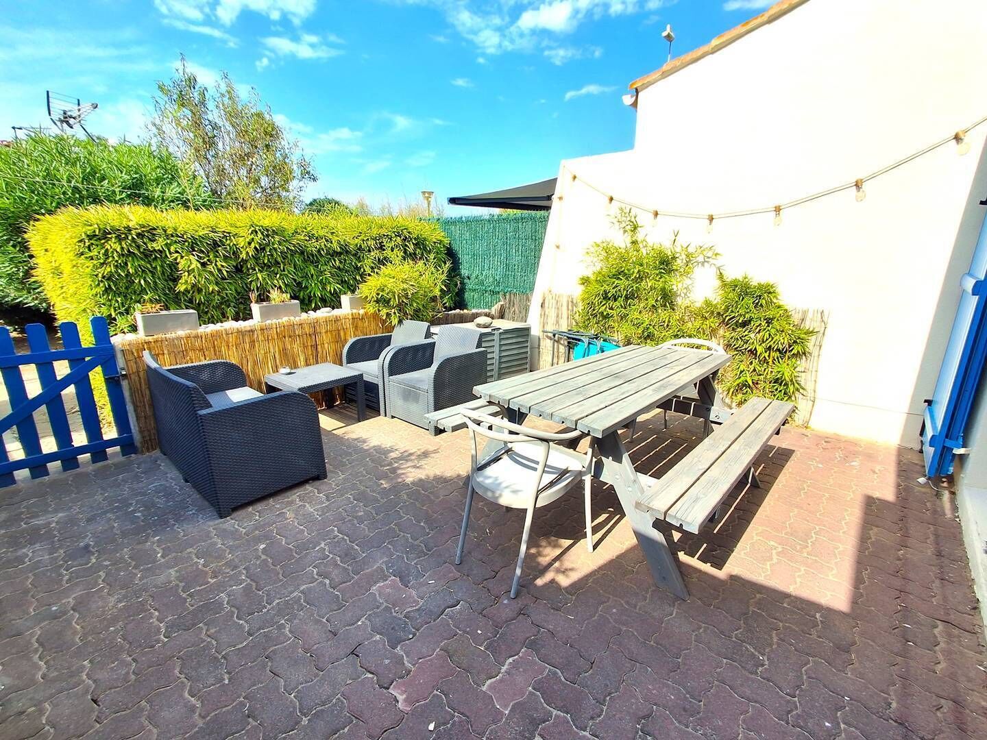 Een terras buiten met rieten meubels, een houten tafel en een blauwe omheining.