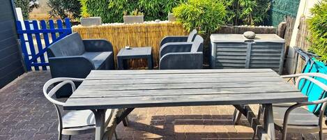 An outdoor patio area with a wooden table and chairs, a blue fence, and various plants.