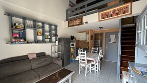 A living room with a grey sofa, a coffee table, a dining table with chairs, a kitchen area, and a staircase leading to an upper level. - STUDIO MEZZANINE POUR 4 PERSONNES CLIMATISE (LE CAP D AGDE)