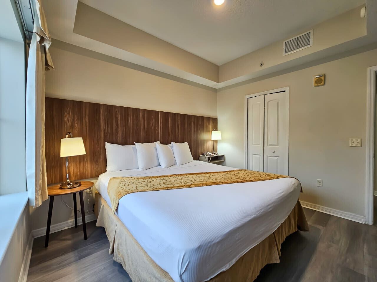 Habitación de hotel con una cama grande, dos mesitas de noche, cabecera de madera, ventana con cortinas y una puerta.