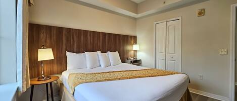 Habitación de hotel con una cama grande, dos mesitas de noche, cabecera de madera, ventana con cortinas y una puerta.