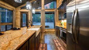 Une cuisine moderne dotée de plans de travail en granit, d’appareils électroménagers en acier inoxydable et de grandes fenêtres offrant une vue sur une zone boisée.