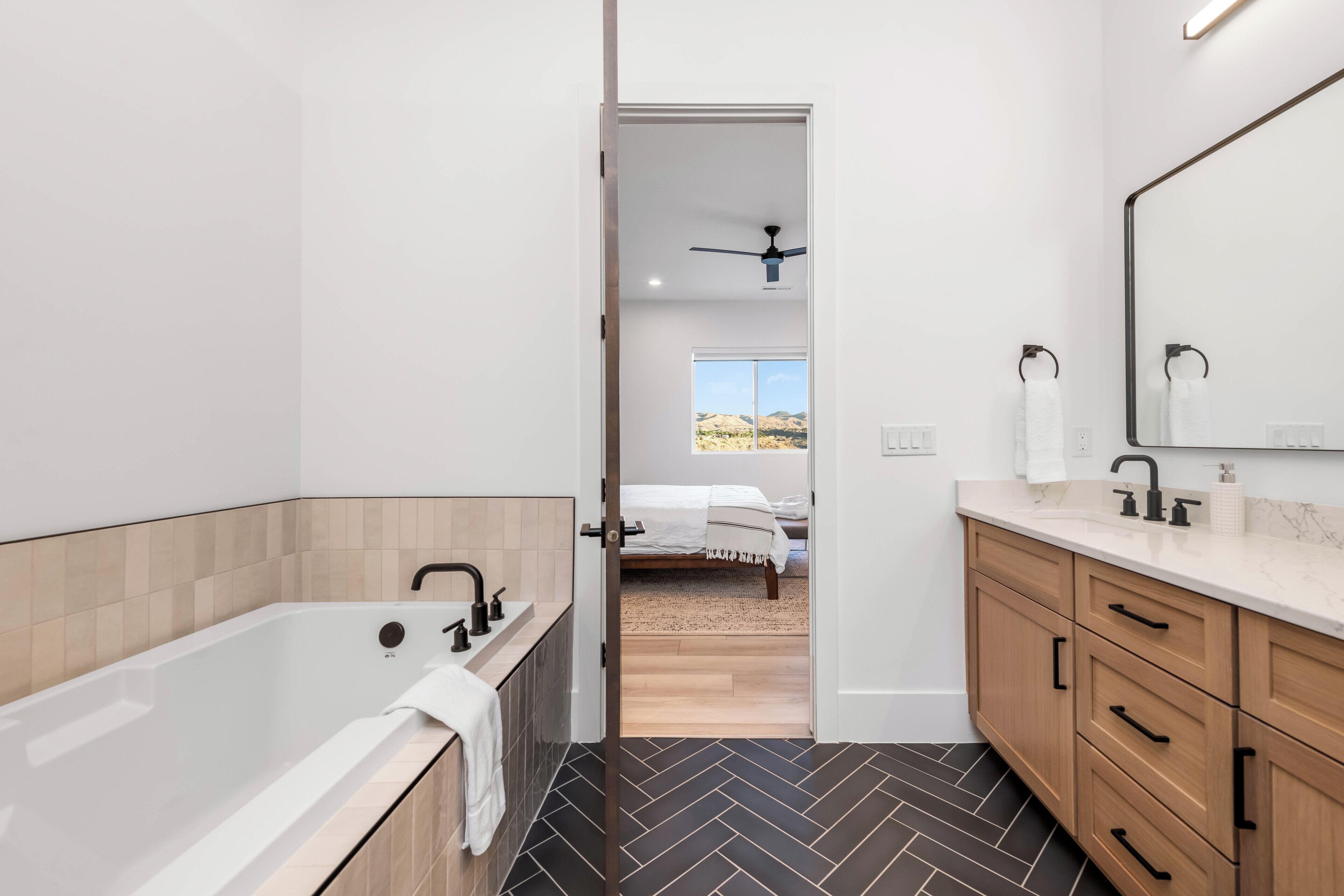 Un baño moderno con bañera, ducha y tocador. Se ve una cama a través de una puerta abierta en la habitación contigua.