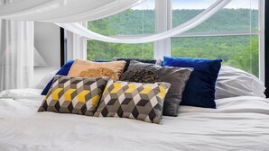 A bed with multiple pillows and a view of a forest through a window. - 8-Person HotTub, Family&Group Friendly, EV Charger, Free Platinum Pool Pass (Mc Gaheysville)
