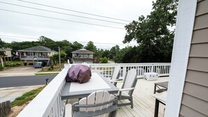 Une terrasse en bois avec une table et des chaises, donnant sur une rue résidentielle bordée de maisons et de voitures garées.
