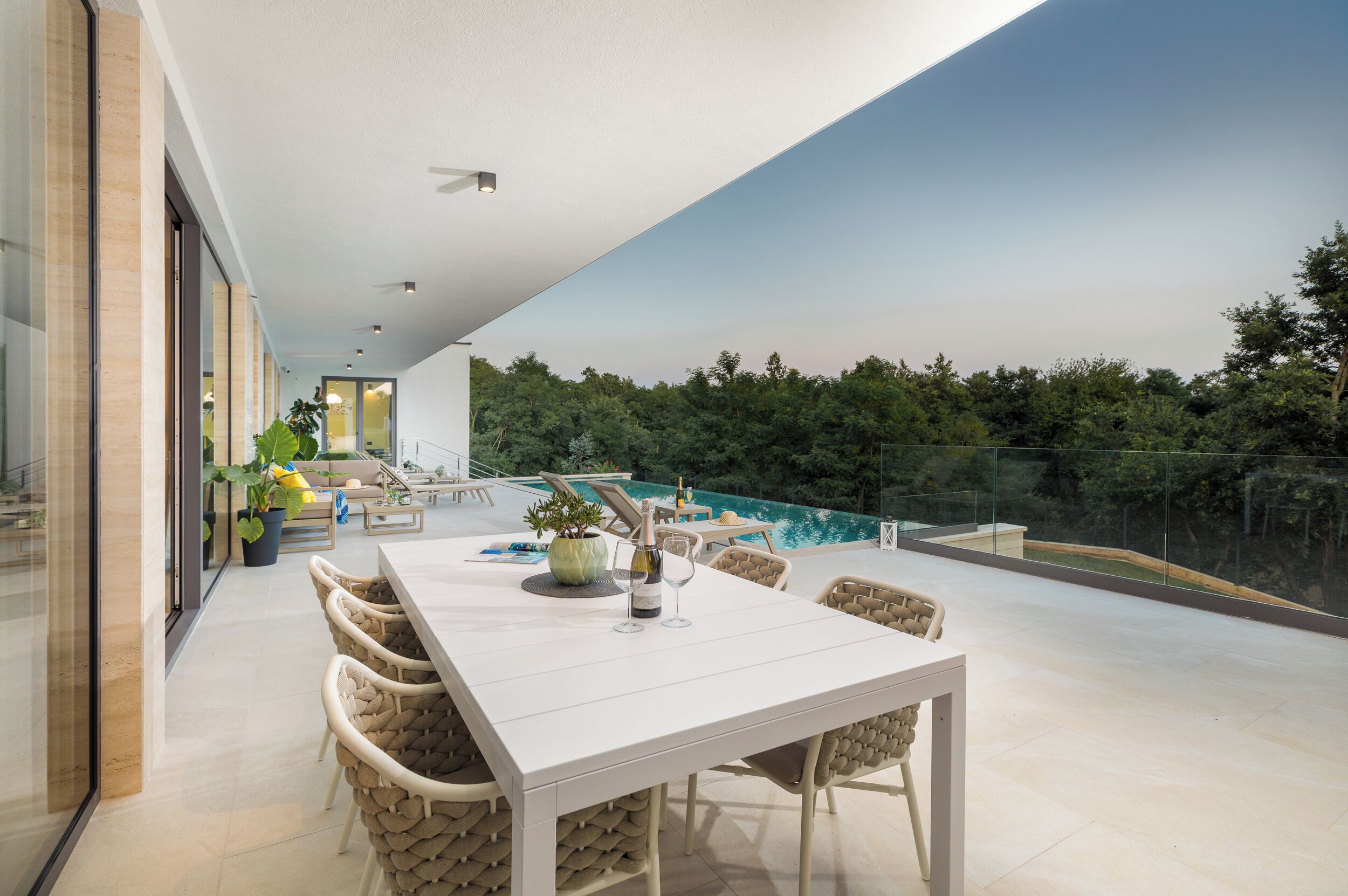 Un área de comedor exterior moderna con una mesa blanca y sillas de mimbre, con vistas a una piscina y abundante vegetación.