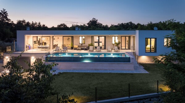 A modern house with a swimming pool, a patio with outdoor furniture, and a well-lit interior.