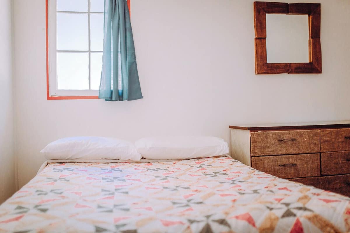 A bedroom with a bed, a wooden dresser, a window with curtains, and a mirror on the wall.