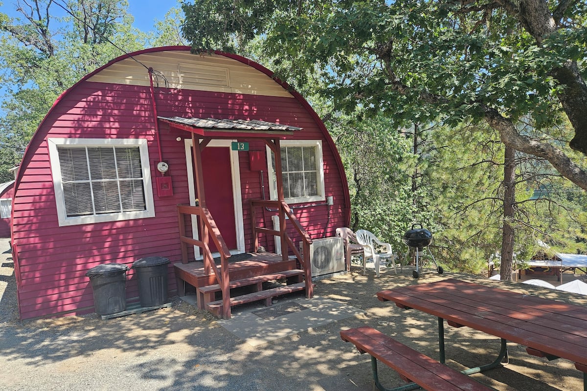 A red wooden cabin with a porch, a picnic table, and a grill.