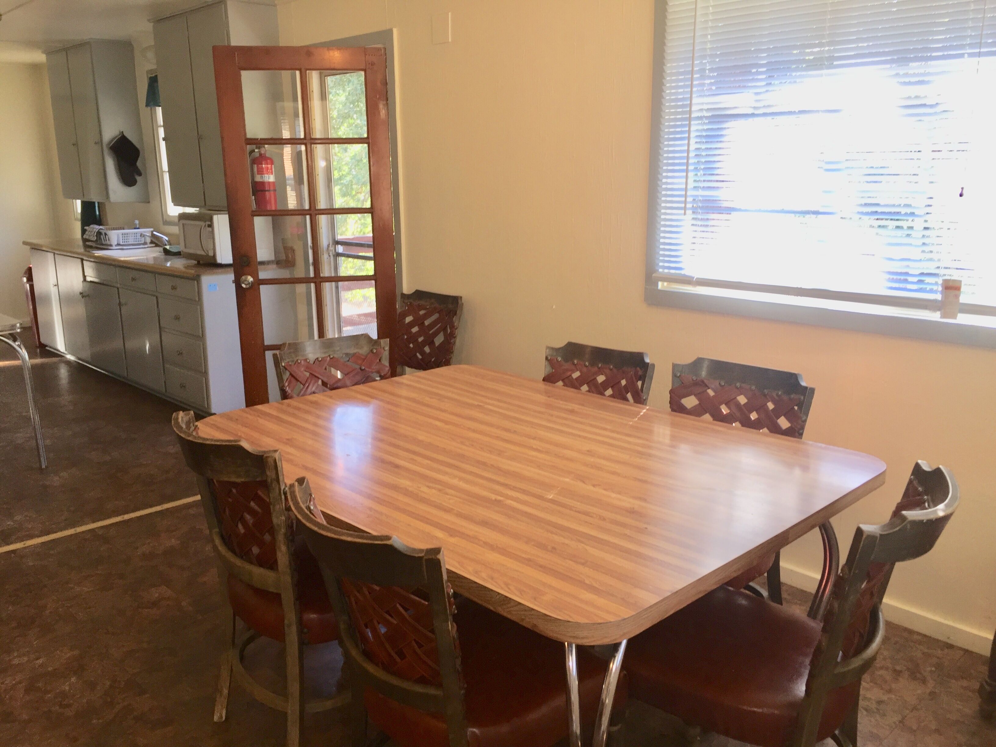 A dining area with a wooden table and six chairs, a kitchen in the background, and a window with blinds.