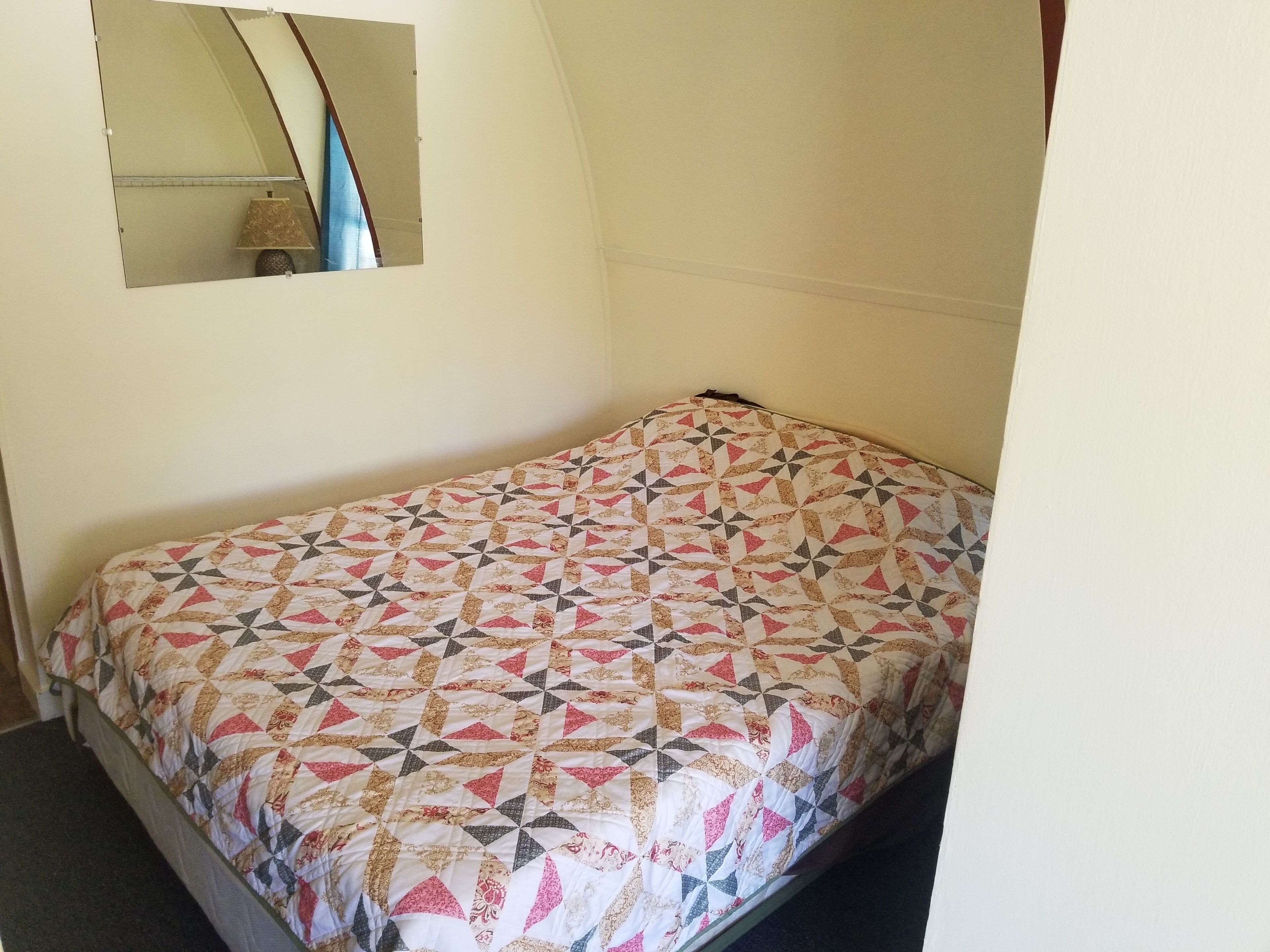A small, single-bed room with a geometric patterned bedspread, a small mirror on the wall, and a lamp on the floor.