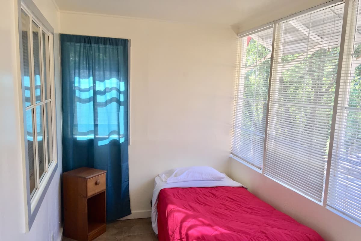 A small bedroom with a single bed, a wooden nightstand, a window with blinds, and a curtain.