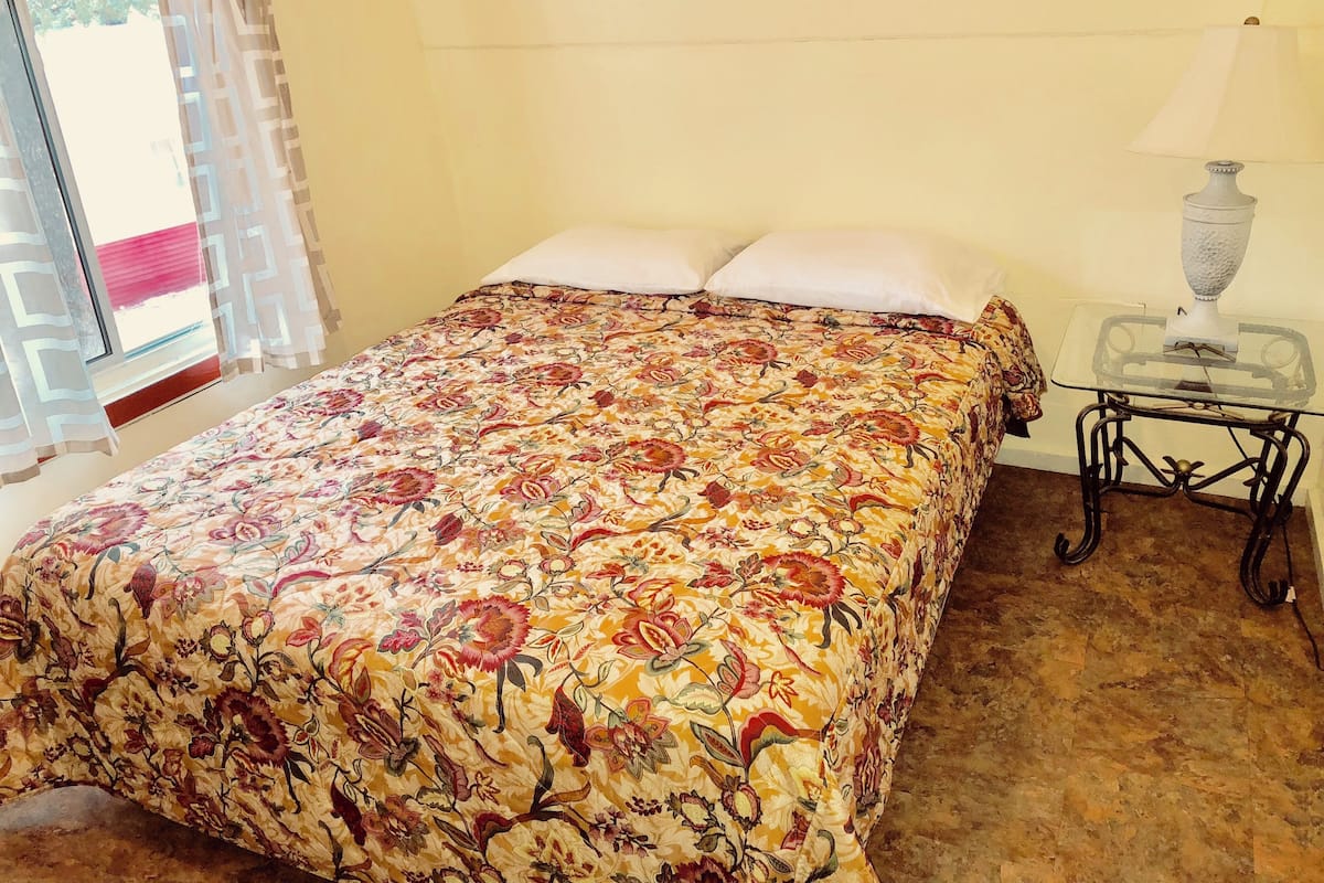 A bed with a floral bedspread, a bedside table with a lamp, and a view of the outdoors through a window with curtains.