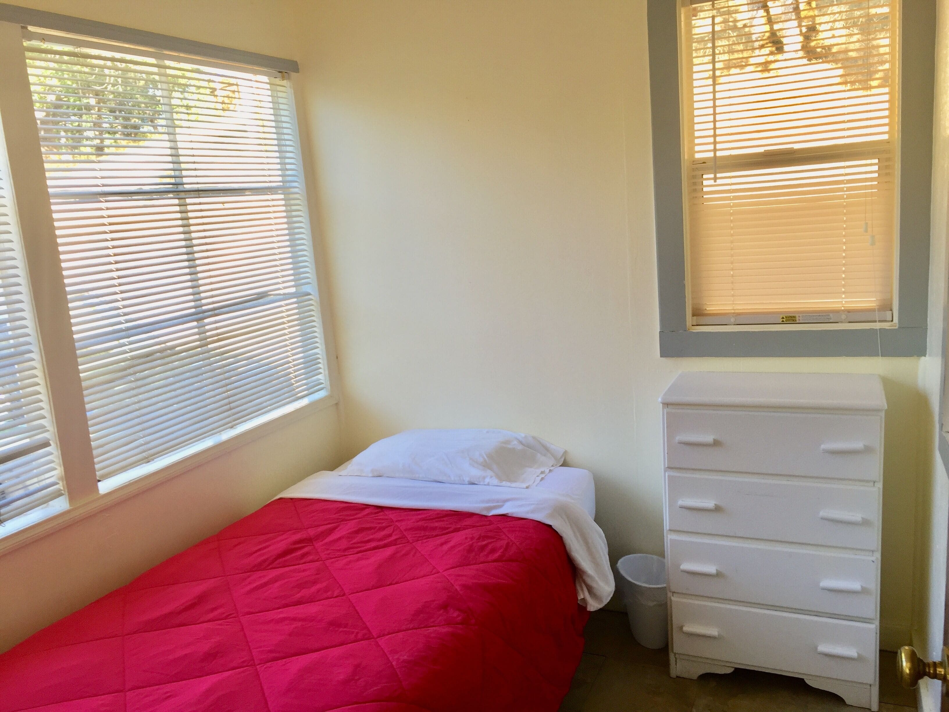 A small, single-bed room with a red bedspread, white bedding, a white dresser, and two windows with blinds.