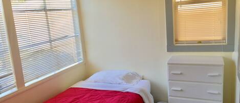 A small, single-bed room with a red bedspread, white bedding, a white dresser, and two windows with blinds.