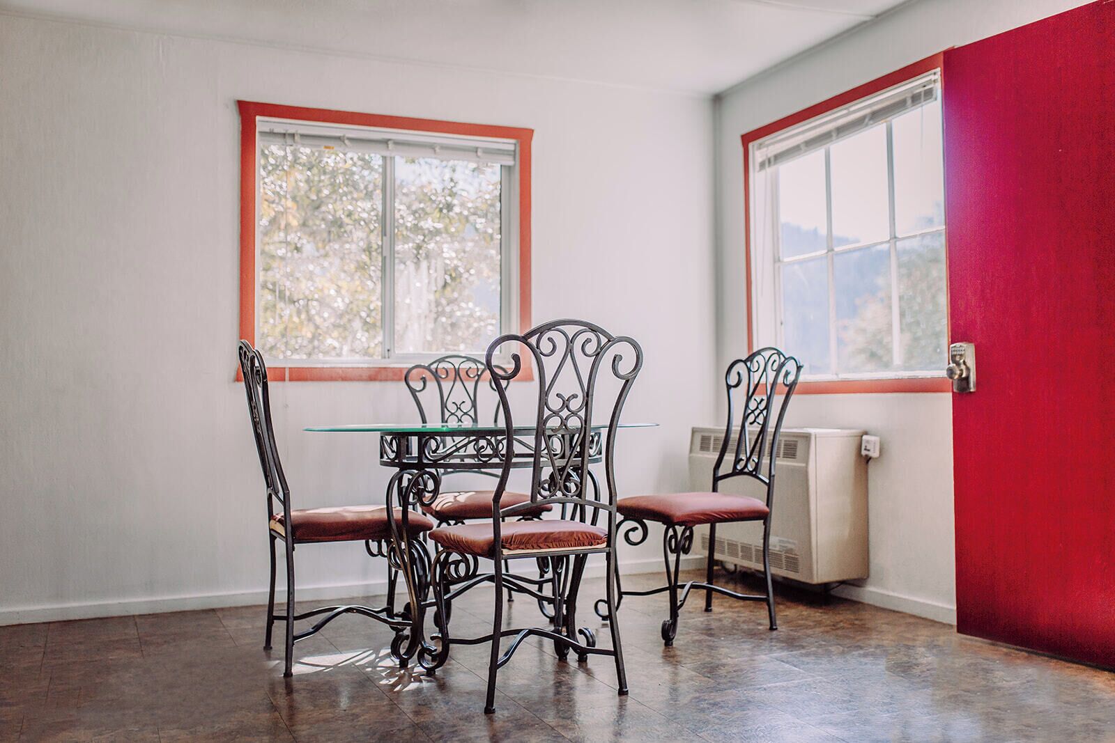 A dining area with a glass table and four chairs, two windows, and a red wall.
