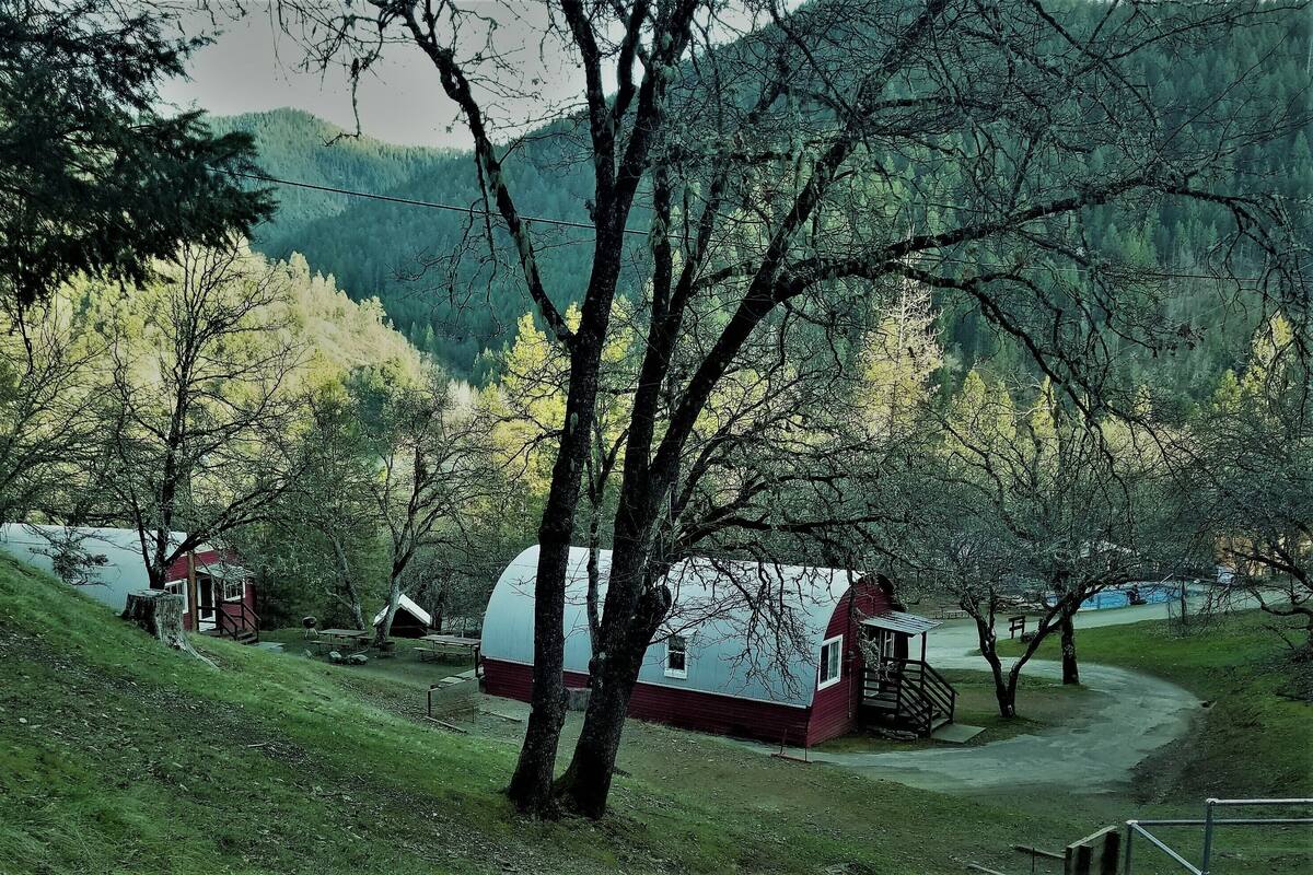 A rustic cabin with a red roof and white walls, surrounded by trees and a lake.