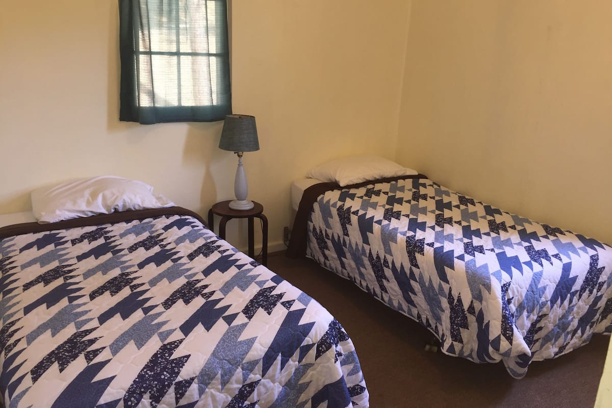 Two single beds with patterned bedding, a bedside table with a lamp, and a window with a curtain.