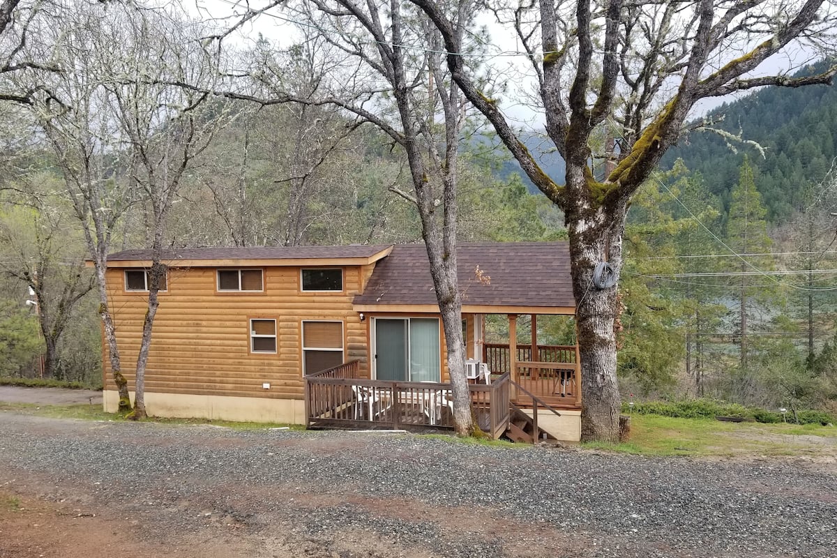 A log cabin with a deck and a large window, surrounded by trees and a gravel driveway.