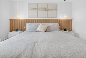 A modern bedroom with a large bed, wooden headboard, bedside tables with plants, and a wall-mounted artwork.