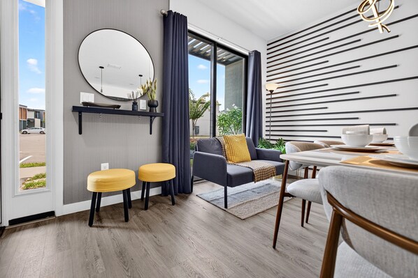 A modern interior with a dining area, a sofa with a yellow cushion, a round mirror, and a view through a glass door.
