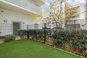 Un patio cercado con césped artificial, un árbol de hojas amarillas y un edificio con fachada blanca.