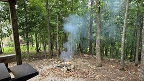Une zone boisée avec un foyer et de la fumée qui s’en échappe, entourée d’arbres et d’une structure en bois.