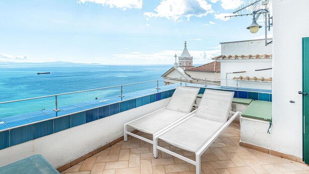 A balcony with a view of the sea, a white lounge chair, and a tiled floor.